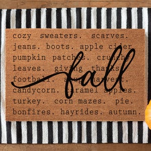 May include: A rectangular doormat with a black and white striped border and a brown center. The center features the word "fall" in cursive, surrounded by autumn-themed words like "sweaters," "pumpkin patches," and "turkey." An orange pumpkin sits to the right.