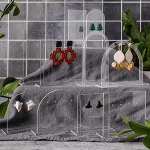 May include: Clear acrylic earring display stands with various pairs of earrings, including a pair of red and black cross-shaped earrings, a pair of green Christmas tree earrings, a pair of white ghost earrings, a pair of black witch hat earrings, and a pair of white and gold dangle earrings.