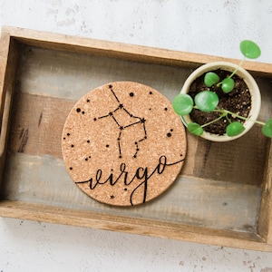 May include: A cork coaster with a black and white design of the Virgo constellation and the word "Virgo" written in cursive. The coaster is on a wooden tray with a small potted plant.