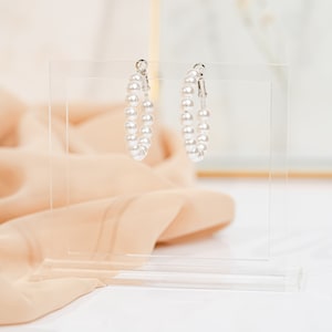 May include: A pair of silver hoop earrings with white pearl accents. The earrings are displayed on a clear acrylic stand.