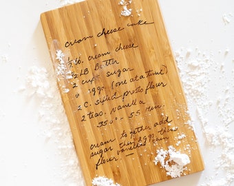 May include: A wooden cutting board with a handwritten recipe for cream cheese cake. The recipe includes ingredients like cream cheese, butter, sugar, eggs, flour, and vanilla. The board is covered in a light dusting of powdered sugar.