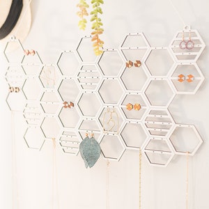 May include: White hexagonal earring holder with various earrings hanging from it. The holder is made of wood and has a honeycomb design.