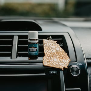 May include: A small bottle of essential oil with a white cap and a blue label that says "Northern Lights Black Spruce" is placed on a black car air vent. A wooden car air freshener shaped like the state of Georgia is hanging from the vent.