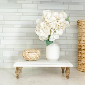 May include: White ceramic vase with a bouquet of white hydrangeas on a white tiered stand with wooden legs. A small woven basket sits on the top tier of the stand.
