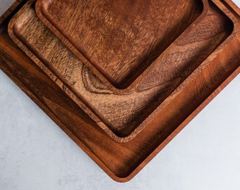 May include: Three square wooden serving trays, nested together, with a light brown wood grain. The trays are made of natural wood and have a rustic look.