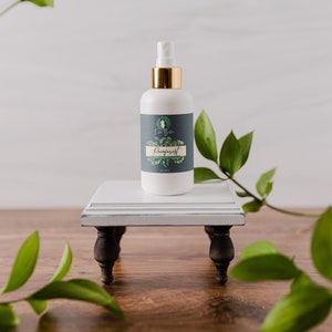 May include: A white spray bottle with a gold cap and a label that reads "Rainforest" sits on a small, white, wooden stand with dark legs. Green leaves and branches frame the bottle against a light background.