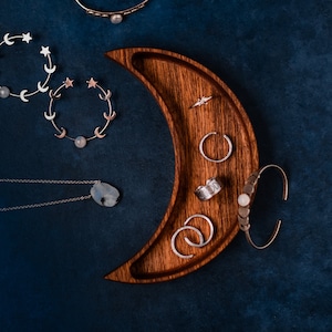 Peut inclure: Un plateau à bijoux en bois en forme de croissant de lune avec des anneaux, des bracelets et des colliers en argent. Le plateau est sur un fond bleu foncé.