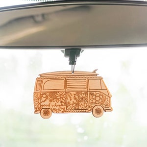 May include: A wooden car air freshener shaped like a vintage van with a surfboard on top. The van is decorated with flowers and the words "good vibes".