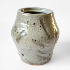 May include: A handmade, ceramic vase with a unique, organic shape. The vase is a muted gray-green color with darker speckles and textured markings. The interior is dark, and the base has a natural, unglazed finish. A decorative piece for the home.