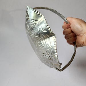 Puede incluir: Una fuente de metal plateado en relieve con asa. El plato tiene un dise&ntilde;o floral y un borde festoneado. El asa es una banda de metal retorcido. El plato se sostiene sobre un fondo gris claro liso.