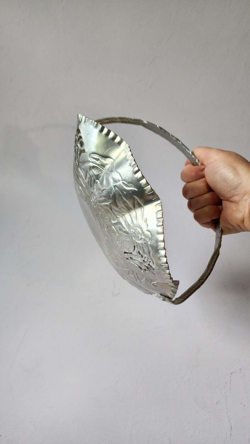 Puede incluir: Una fuente de metal plateado en relieve con asa. El plato tiene un dise&ntilde;o floral y un borde festoneado. El asa es una banda de metal retorcido. El plato se sostiene sobre un fondo gris claro liso.