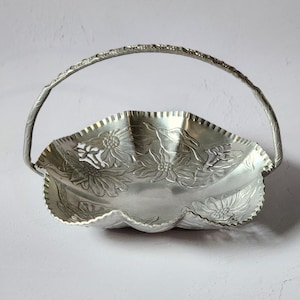 Puede incluir: Cesta de metal plateado con asa. La cesta tiene un borde festoneado y est&aacute; decorada con dise&ntilde;os florales en relieve. El asa tambi&eacute;n es plateada y tiene un patr&oacute;n texturizado. La cesta probablemente se utiliza para servir o exhibir.