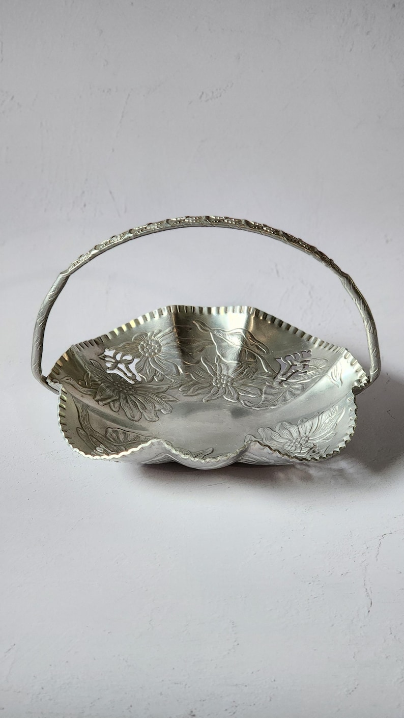 Puede incluir: Cesta de metal plateado con asa. La cesta tiene un borde festoneado y est&aacute; decorada con dise&ntilde;os florales en relieve. El asa tambi&eacute;n es plateada y tiene un patr&oacute;n texturizado. La cesta probablemente se utiliza para servir o exhibir.
