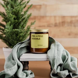 Może przedstawiać: Świeca zapachowa Fir + Pine Needle w szklanym słoiku ze złotą pokrywką, stojąca na białym i szarym dekoracyjnym stojaku. Na zdjęciu znajduje się r&oacute;wnież małe drzewko iglaste w doniczce i szałwiowa zielona szmatka. Etykieta świecy zawiera tekst "FIR + PINE NEEDLE".