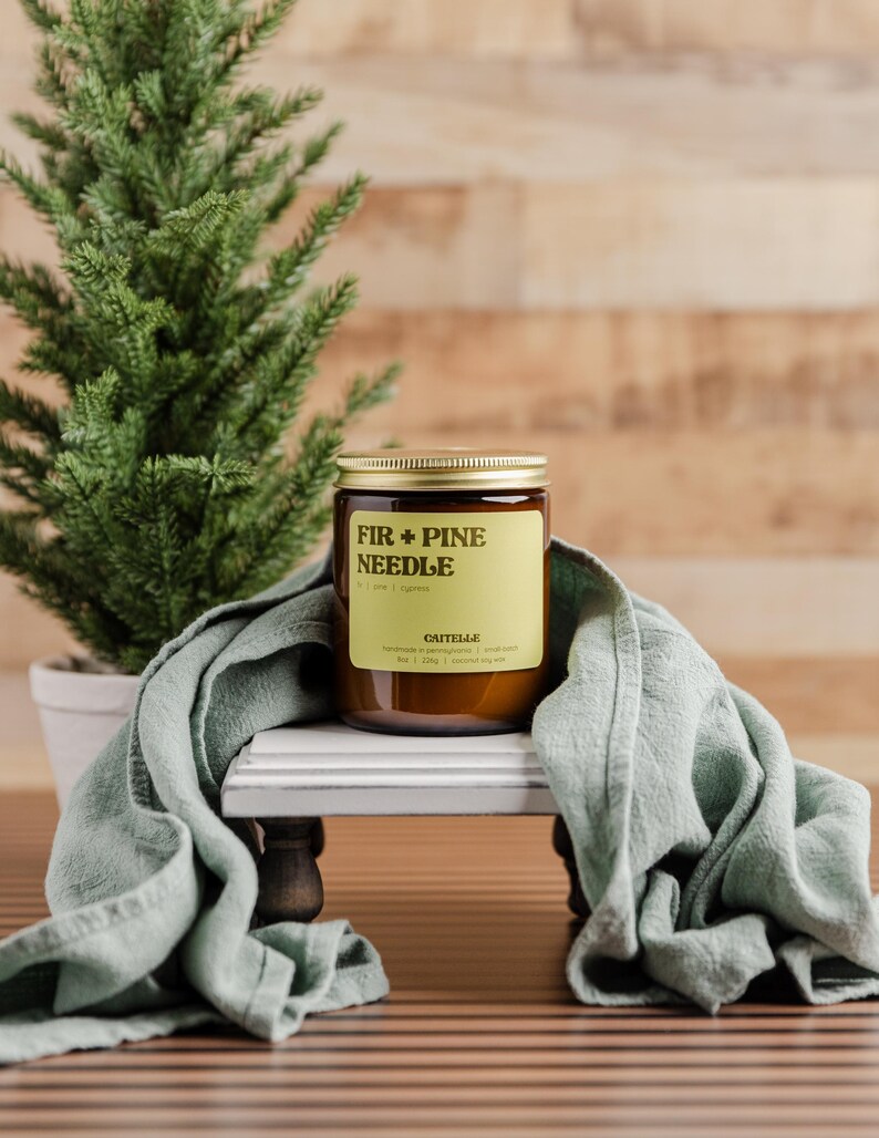 Może przedstawiać: Świeca zapachowa Fir + Pine Needle w szklanym słoiku ze złotą pokrywką, stojąca na białym i szarym dekoracyjnym stojaku. Na zdjęciu znajduje się r&oacute;wnież małe drzewko iglaste w doniczce i szałwiowa zielona szmatka. Etykieta świecy zawiera tekst "FIR + PINE NEEDLE".