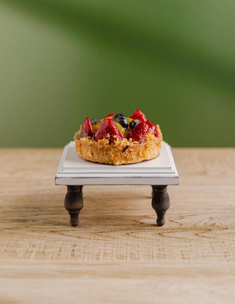 Może przedstawiać: Miniaturowa tarta owocowa z truskawkami, jagodami i innymi owocami stoi na małym, białym, dekoracyjnym stoliku z ciemnobrązowymi n&oacute;żkami. Tarta ma złoto-brązowy sp&oacute;d i kolorowy asortyment świeżych owoc&oacute;w. St&oacute;ł stoi na drewnianej powierzchni.