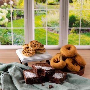 Może przedstawiać: Asortyment wypiek&oacute;w, w tym czekoladowe brownie posypane cukrem pudrem, ciasteczka z kawałkami czekolady i pączki. Smakołyki ułożone są na drewnianej powierzchni z zieloną serwetą i białym stojakiem. W tle okno z widokiem na ogr&oacute;d.