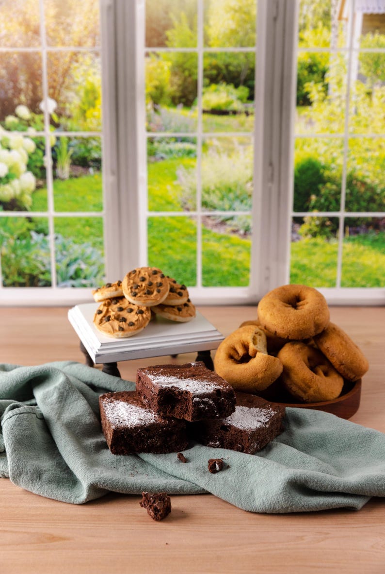 Może przedstawiać: Asortyment wypiek&oacute;w, w tym czekoladowe brownie posypane cukrem pudrem, ciasteczka z kawałkami czekolady i pączki. Smakołyki ułożone są na drewnianej powierzchni z zieloną serwetą i białym stojakiem. W tle okno z widokiem na ogr&oacute;d.