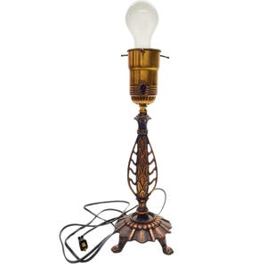 May include: A vintage table lamp with a bronze finish. The lamp features a decorative base, a detailed stem, and a brass-colored bulb socket. The lamp is topped with a standard light bulb and has a black power cord.