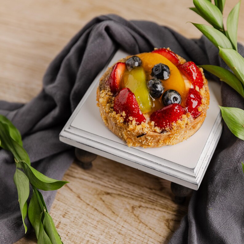 Może przedstawiać: Tarta owocowa z truskawkami, jagodami, plasterkami brzoskwini i kiwi na białej drewnianej tacy. Tarta ma kruszonkę, z szarą szmatką i zielonymi liśćmi.