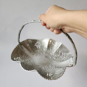 Puede incluir: Una cesta plateada con borde festoneado y asa, sostenida por una mano. La cesta presenta dise&ntilde;os florales en relieve. El asa est&aacute; torcida y tiene un patr&oacute;n floral. La cesta es de metal.