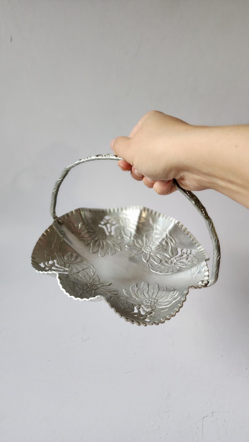 Puede incluir: Una cesta plateada con borde festoneado y asa, sostenida por una mano. La cesta presenta dise&ntilde;os florales en relieve. El asa est&aacute; torcida y tiene un patr&oacute;n floral. La cesta es de metal.