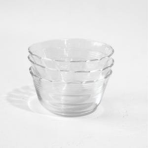 May include: A stack of three clear glass bowls with a scalloped rim design. Each bowl features a smooth, transparent surface with subtle horizontal ridges along the exterior walls, creating a uniform and simple appearance for these kitchen vessels.