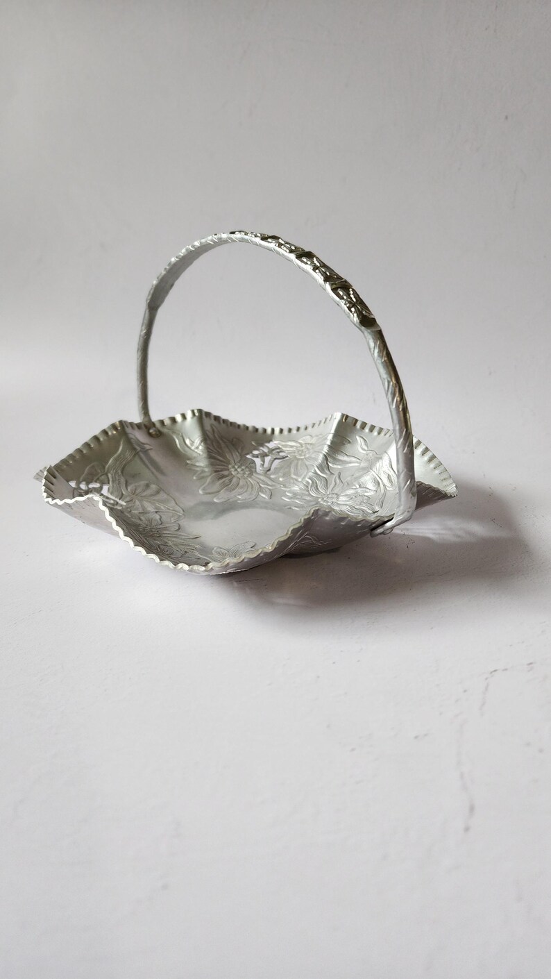 Puede incluir: Una cesta de metal plateado con asa. La cesta tiene un borde festoneado y est&aacute; decorada con motivos florales. El asa es arqueada y tiene un dise&ntilde;o texturizado. La cesta probablemente se utiliza con fines decorativos.