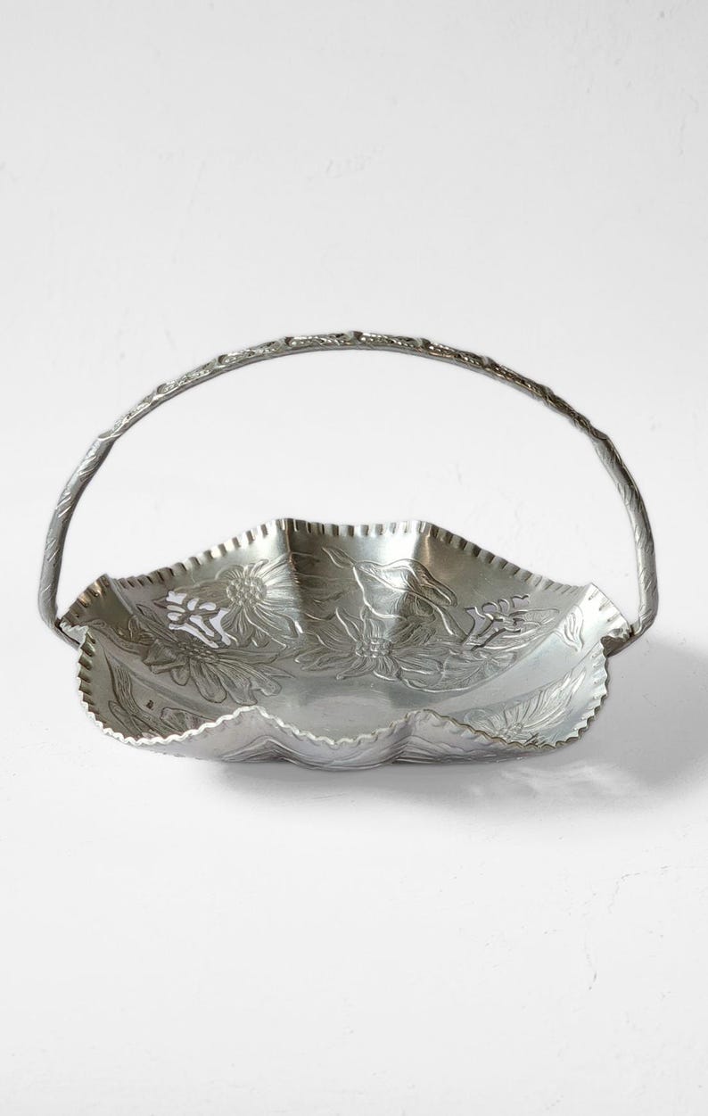Puede incluir: Una cesta de metal plateado con asa. La cesta tiene un borde festoneado y est&aacute; decorada con dise&ntilde;os florales en relieve. El asa arqueada tiene un acabado texturizado, lo que realza el aspecto decorativo de la cesta. Ideal para servir o exhibir.