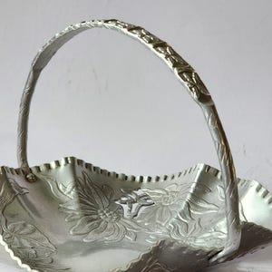 Puede incluir: Una cesta de metal plateado con asa. La cesta tiene un borde festoneado y est&aacute; decorada con dise&ntilde;os florales en relieve. El asa es arqueada y texturizada. La cesta probablemente se utiliza para servir o exhibir.