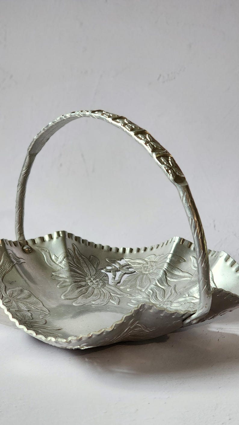 Puede incluir: Una cesta de metal plateado con asa. La cesta tiene un borde festoneado y est&aacute; decorada con dise&ntilde;os florales en relieve. El asa es arqueada y texturizada. La cesta probablemente se utiliza para servir o exhibir.