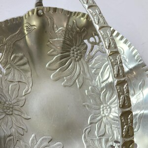 Puede incluir: Cesta de metal plateado con asa. La cesta presenta dise&ntilde;os florales en relieve, incluyendo girasoles, en su superficie. El asa tambi&eacute;n est&aacute; decorada con motivos florales. El borde tiene un borde festoneado.
