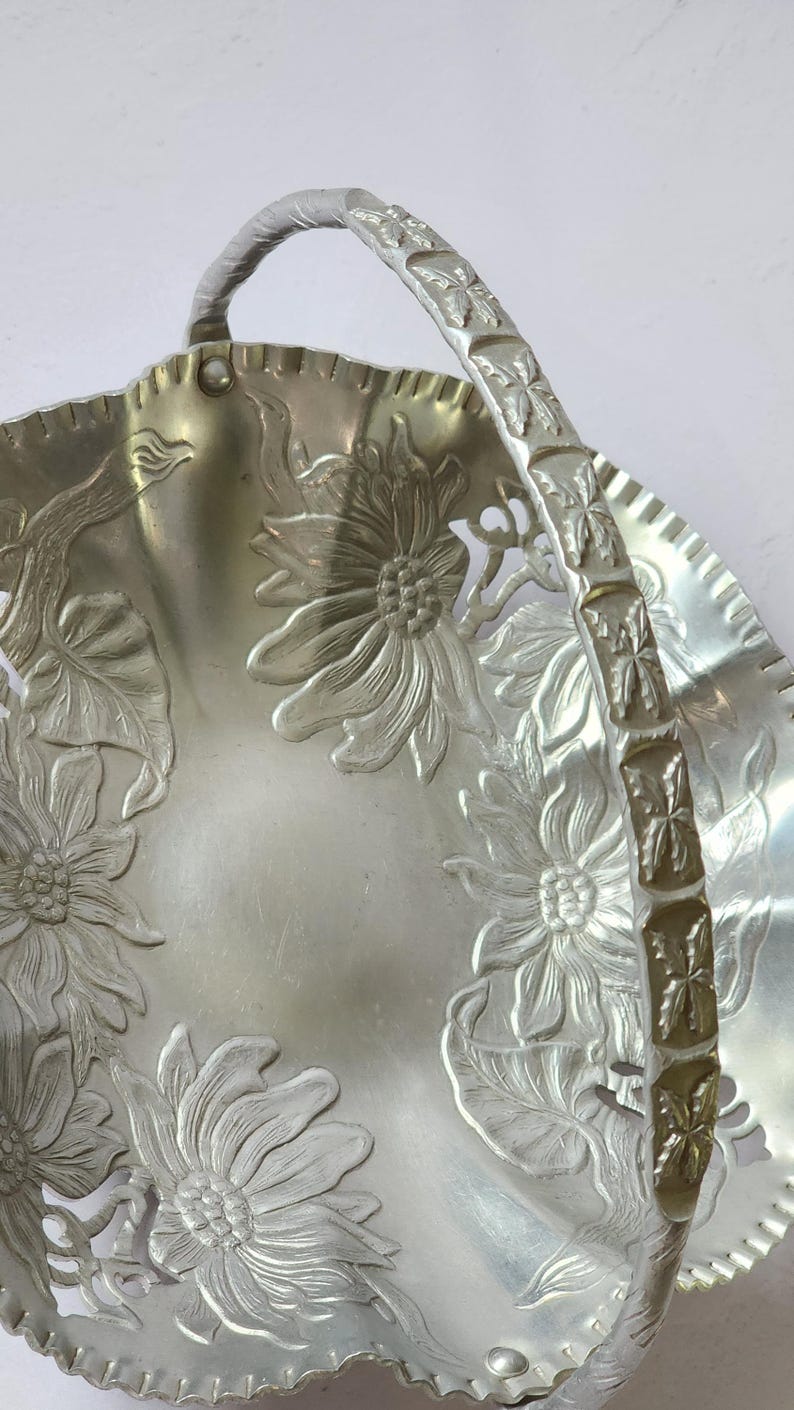 Puede incluir: Cesta de metal plateado con asa. La cesta presenta dise&ntilde;os florales en relieve, incluyendo girasoles, en su superficie. El asa tambi&eacute;n est&aacute; decorada con motivos florales. El borde tiene un borde festoneado.