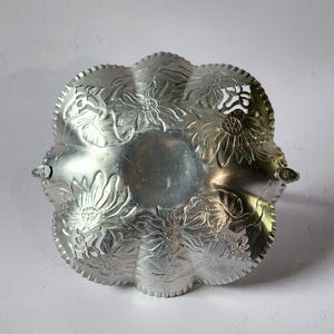 Puede incluir: Un plato para servir de color plateado con un dise&ntilde;o floral. El plato tiene un borde festoneado y est&aacute; decorado con flores en relieve. Dos asas peque&ntilde;as est&aacute;n en lados opuestos. El plato es probablemente de metal y es adecuado para servir alimentos o como pieza decorativa.