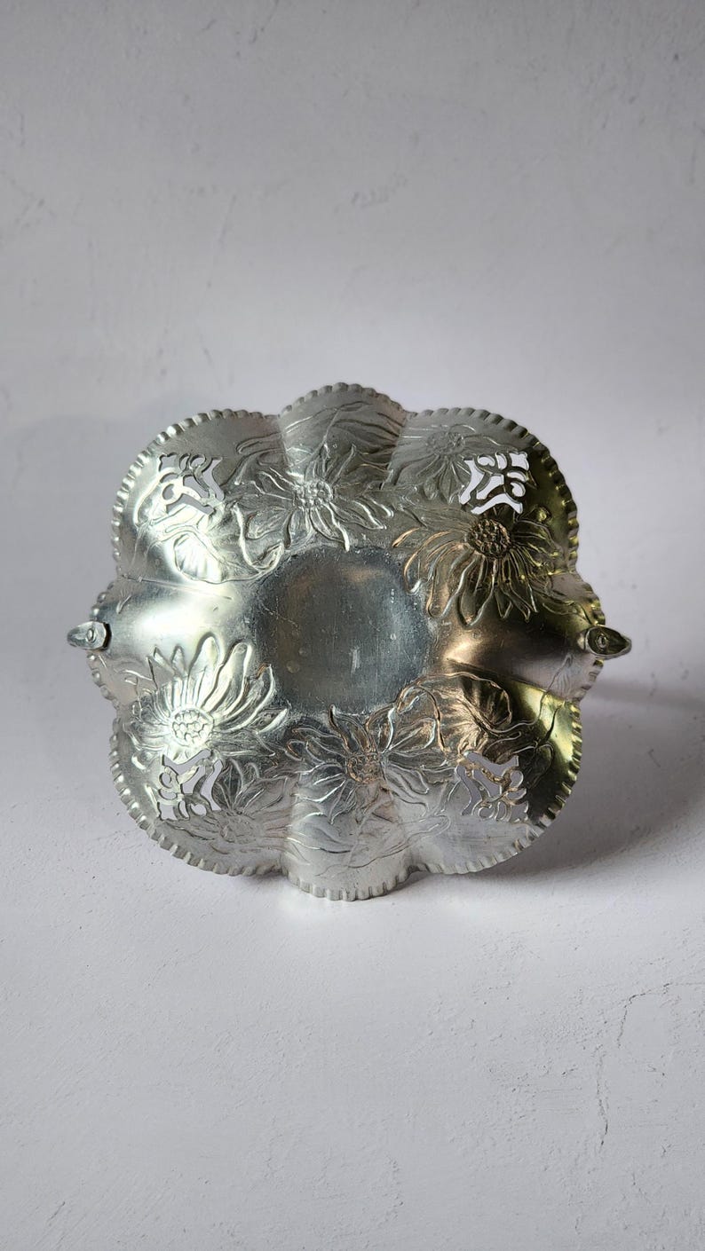 Puede incluir: Un plato para servir de color plateado con un dise&ntilde;o floral. El plato tiene un borde festoneado y est&aacute; decorado con flores en relieve. Dos asas peque&ntilde;as est&aacute;n en lados opuestos. El plato es probablemente de metal y es adecuado para servir alimentos o como pieza decorativa.