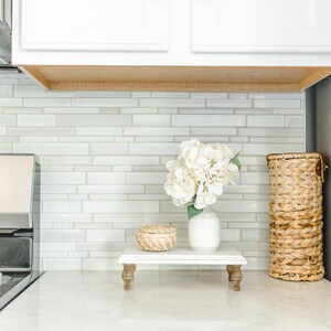 May include: White ceramic vase with white flowers on a white tiered stand with a small woven basket. The stand has four wooden legs. The background is a white tiled wall.