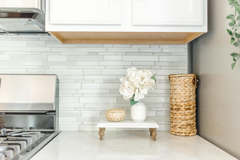 May include: White ceramic vase with white flowers on a white tiered stand with a small woven basket. The stand has four wooden legs. The background is a white tiled wall.