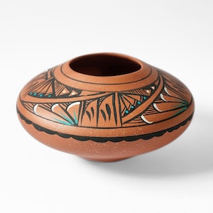 May include: A terracotta-colored ceramic pot with a rounded shape and a small opening. The pot is decorated with black, white, and turquoise geometric patterns. A black scalloped border encircles the base.