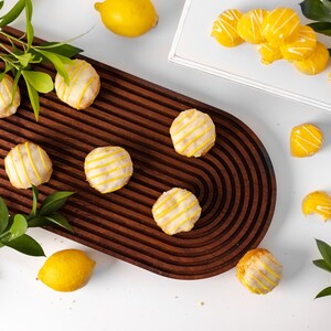 May include: A wooden serving tray with a rounded edge, featuring six lemon-shaped cookies with white icing and yellow stripes. The tray is surrounded by lemons and green leaves.