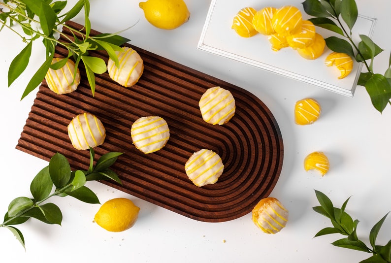 May include: A wooden serving tray with a rounded edge, featuring six lemon-shaped cookies with white icing and yellow stripes. The tray is surrounded by lemons and green leaves.
