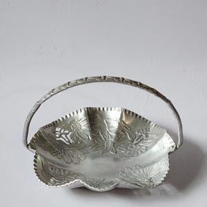 Puede incluir: Cesta de metal plateado con asa. El borde es festoneado y est&aacute; decorado con dise&ntilde;os florales en relieve. El asa tambi&eacute;n es plateada y tiene un patr&oacute;n texturizado. La cesta se utiliza probablemente para servir o exhibir.