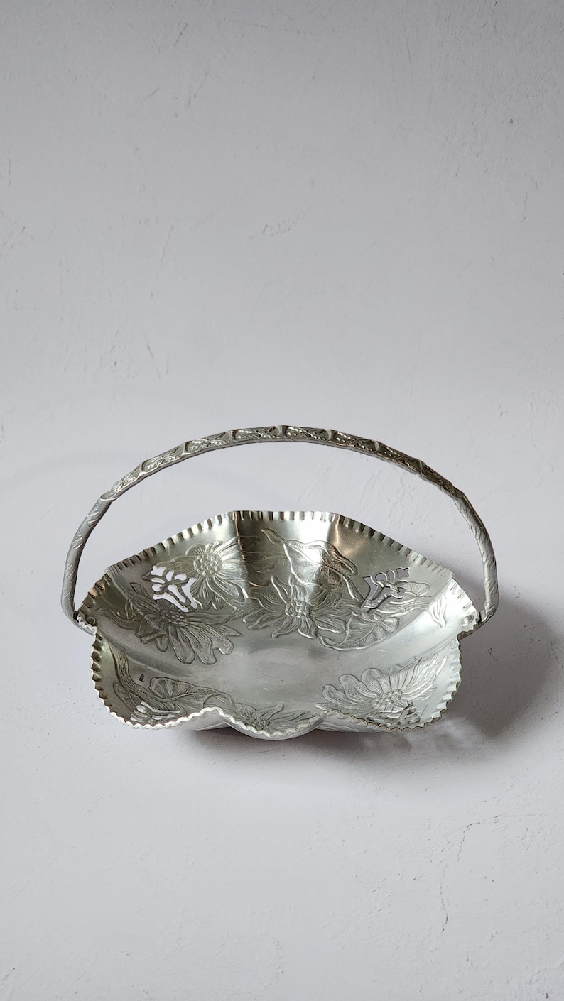 Puede incluir: Cesta de metal plateado con asa. El borde es festoneado y est&aacute; decorado con dise&ntilde;os florales en relieve. El asa tambi&eacute;n es plateada y tiene un patr&oacute;n texturizado. La cesta se utiliza probablemente para servir o exhibir.