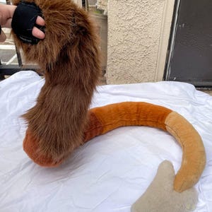 Op de afbeelding: Een pluche, bruine en oranje dierenstaart met een zwarte band. De staart is gemaakt van imitatiebont en heeft een gebogen vorm, met een lichtere punt. De staart is ontworpen om als kostuumaccessoire te worden gedragen.