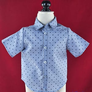 May include: Light blue short-sleeved shirt with a polka dot pattern. The shirt has a button-down front and a classic collar. The shirt is displayed on a mannequin against a red background. The text "@sewniasdollboutique" is visible in the bottom right corner.