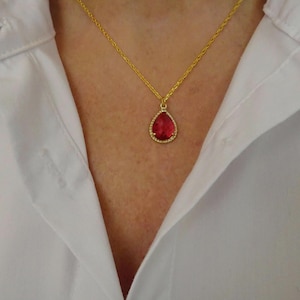May include: A gold chain necklace featuring a teardrop-shaped pendant with a deep red gemstone center surrounded by a thin border of small clear stones. The pendant hangs from a delicate gold-toned metal chain against a white collared shirt.