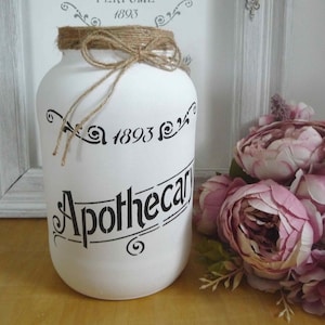 May include: A white apothecary jar with black lettering and a twine bow. The jar is decorated with the words "Apothecary" and "1893". A bouquet of pink flowers sits nearby.