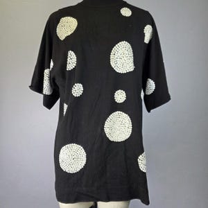 May include: A black short-sleeved shirt featuring a pattern of large, circular white polka dots. The dots are composed of small, densely packed white sequins that create a textured, reflective surface against the solid black fabric of the shirt.