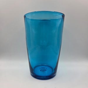 May include: A translucent, vibrant blue glass tumbler with a slightly tapered cylindrical shape. The glass features a smooth, polished surface and a thick, solid base with a visible circular indentation in the center of the bottom interior.