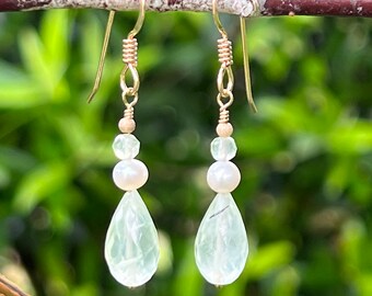 May include: A pair of gold dangle earrings with a teardrop-shaped green gemstone and a white pearl bead on each earring.