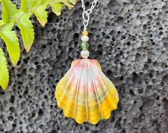 May include: A seashell pendant necklace with a silver chain. The shell is yellow, orange, and white, with a wire wrap. The necklace has a strand of small beads in yellow, green, and white. The background is a dark, textured surface.